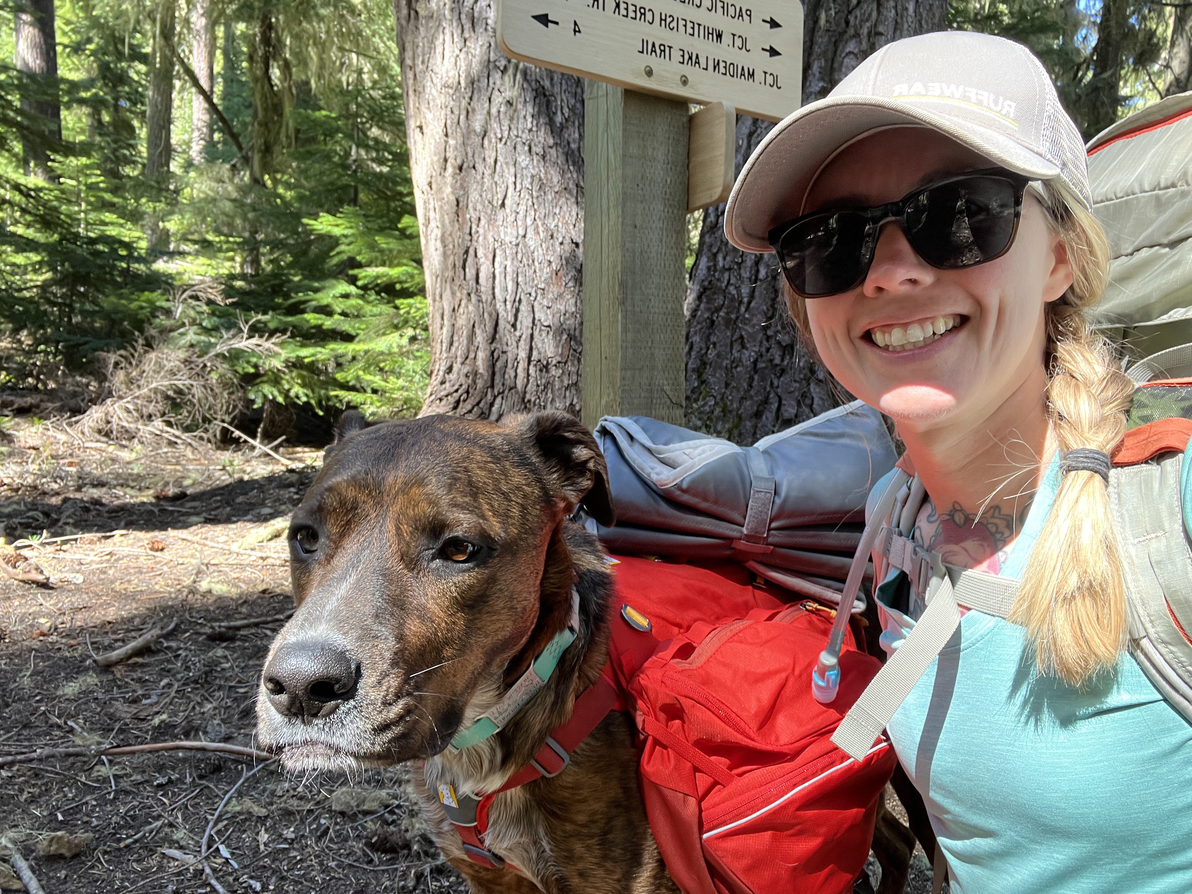 A woman poses with her dog by a hiking trail sign. 