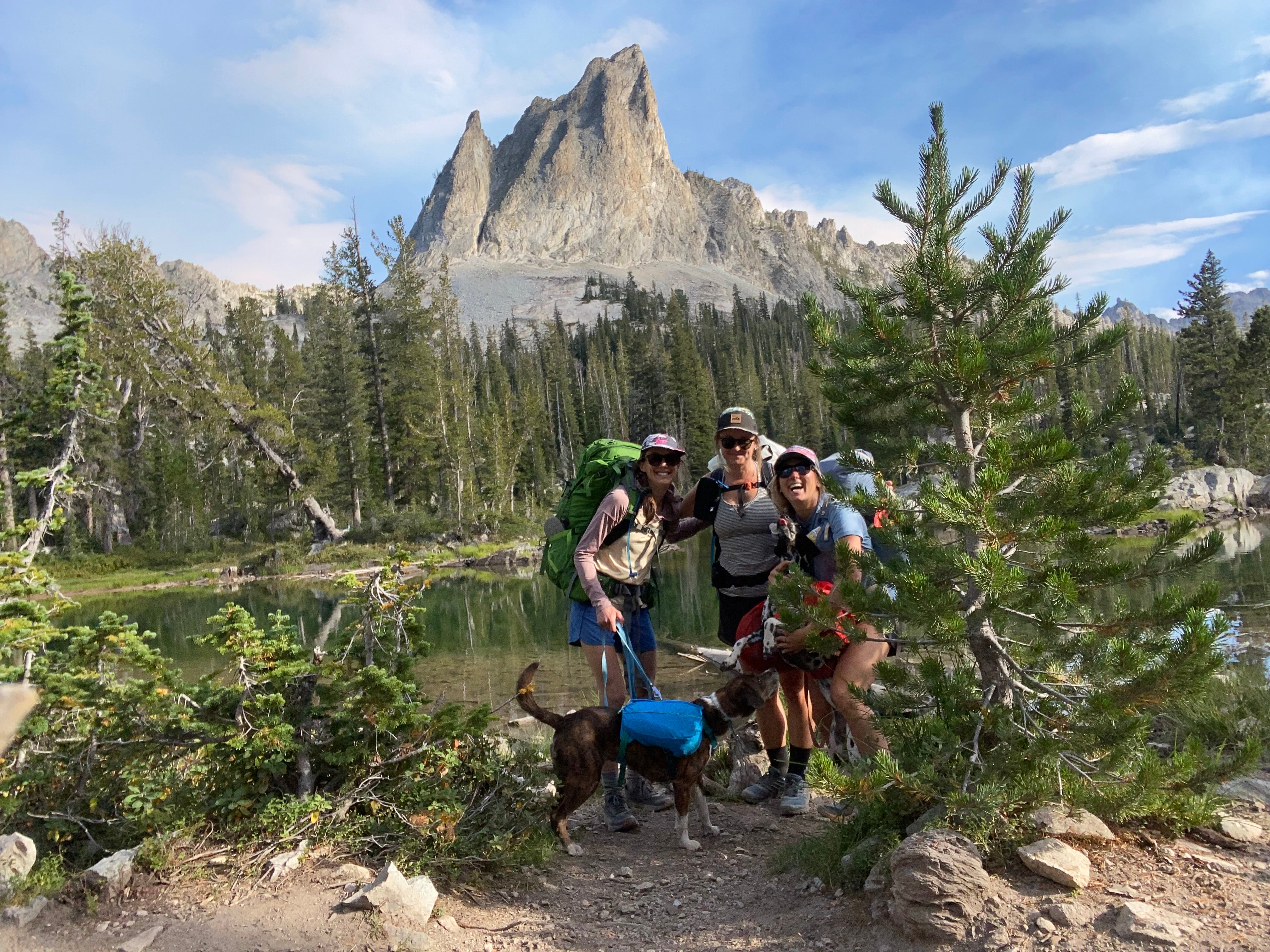 Three women and two dogs backpacking in front of a towering peak