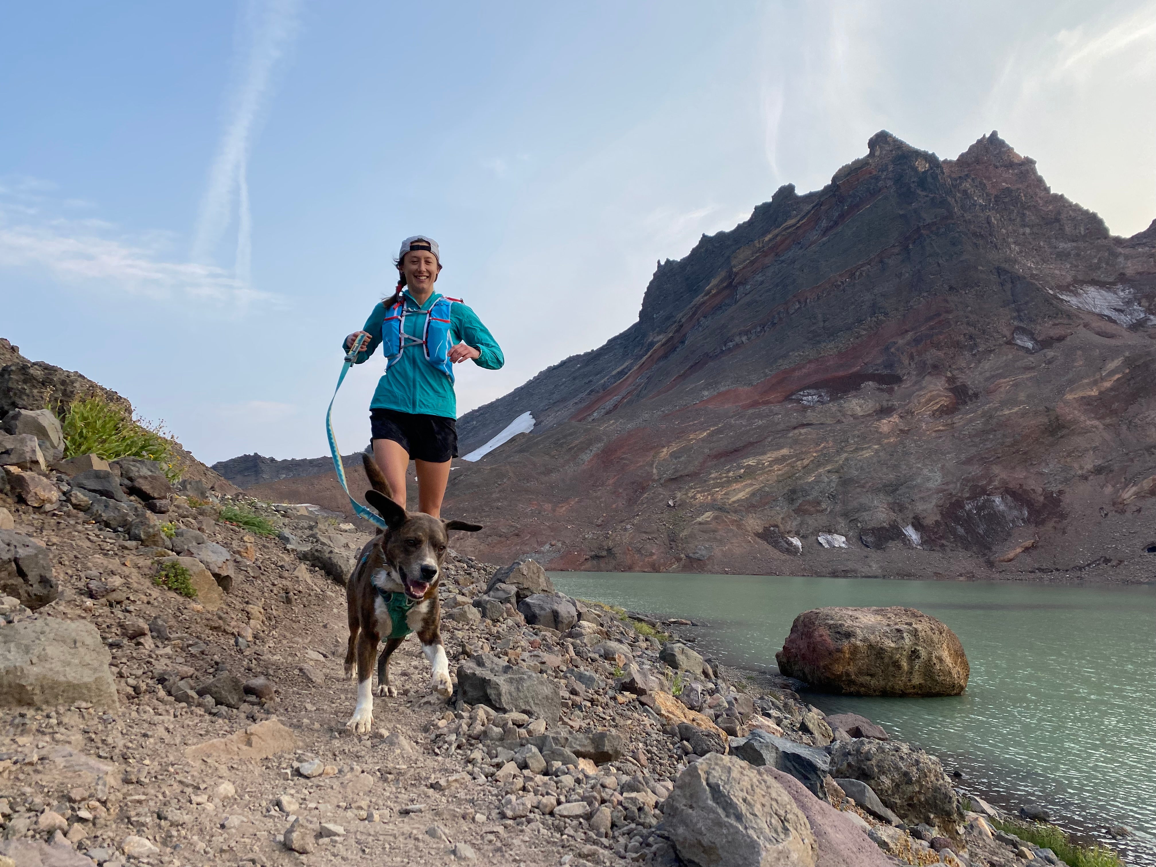 A woman and her dog run on a gravel trail by a mountain lake. 