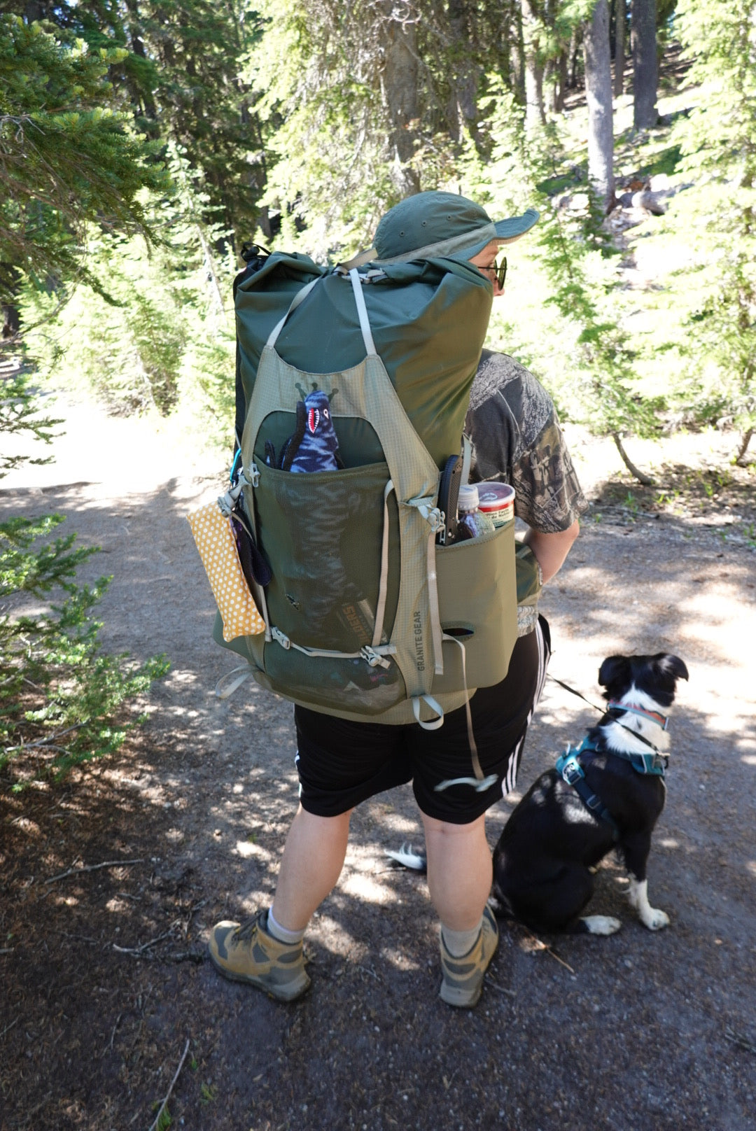 Woman with large backpacking pack stuffed full, with dog in front range harness at her side.