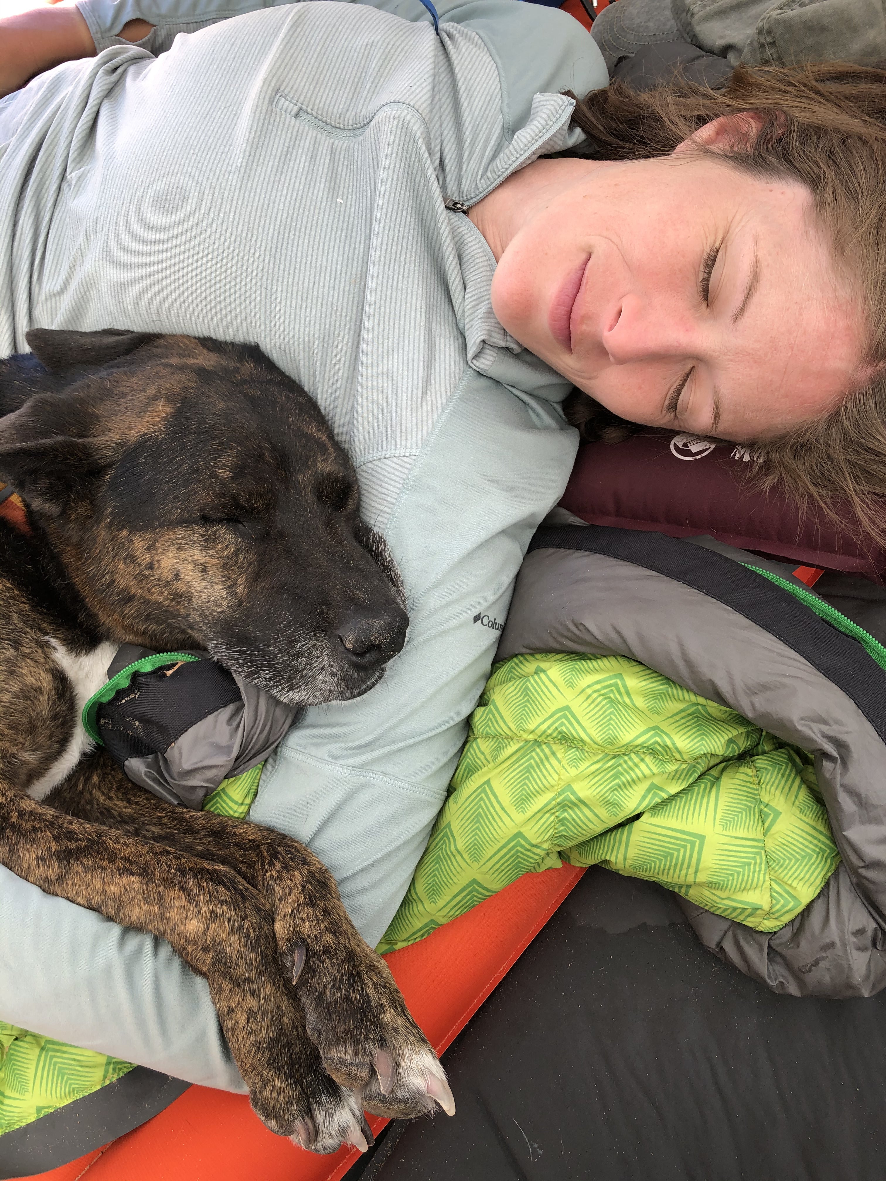 A woman lies down on a sleeping bag with her dog. 