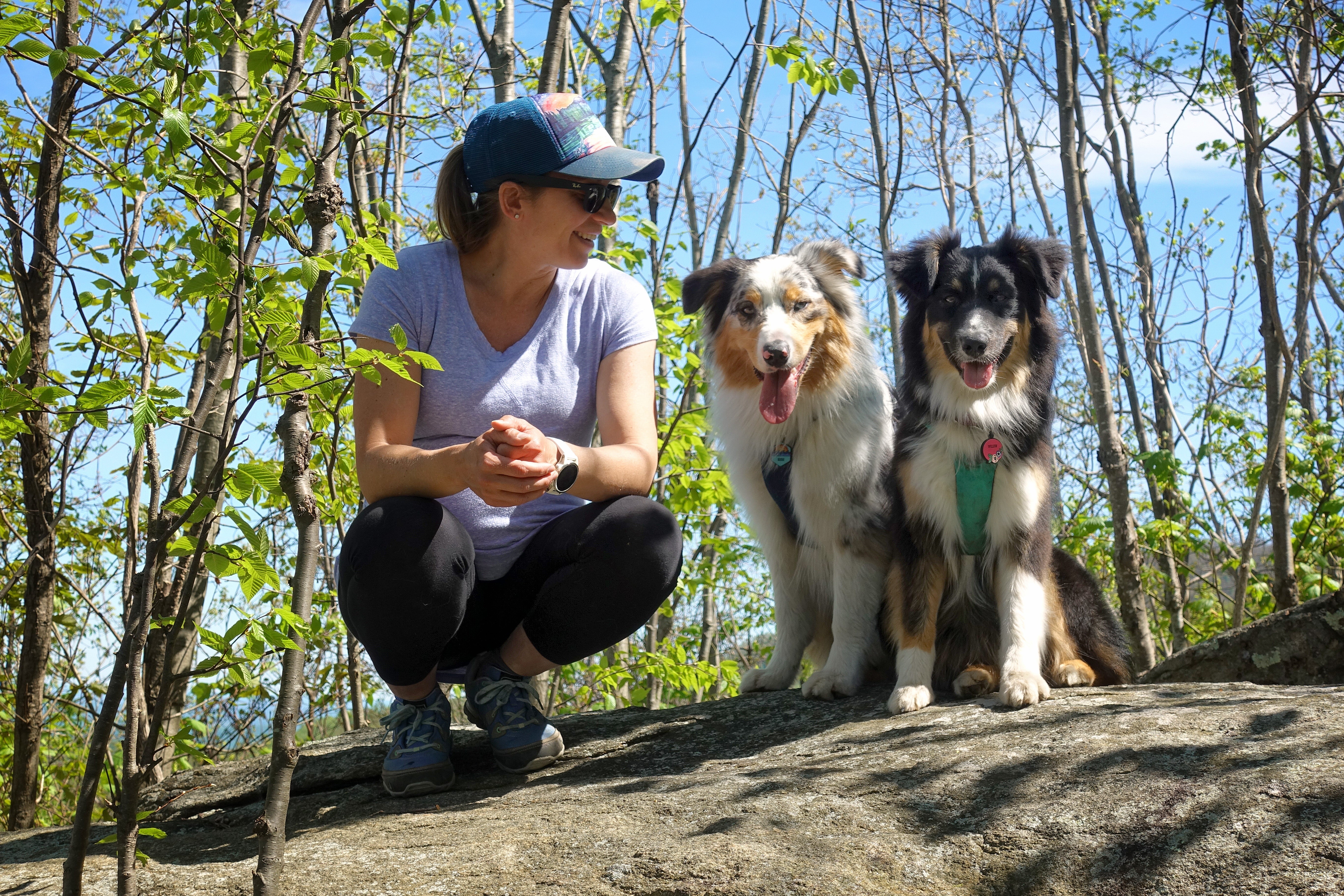 Maria in artist series trucker hat with her two australian shepherds in front range harnesses sitting next to each other on a big rock slab outdoors..