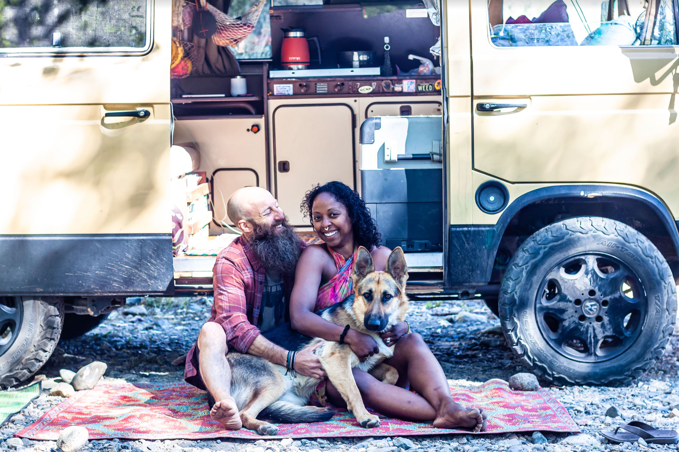 Man, woman, and dog sitting in front of their camper van and smiling.