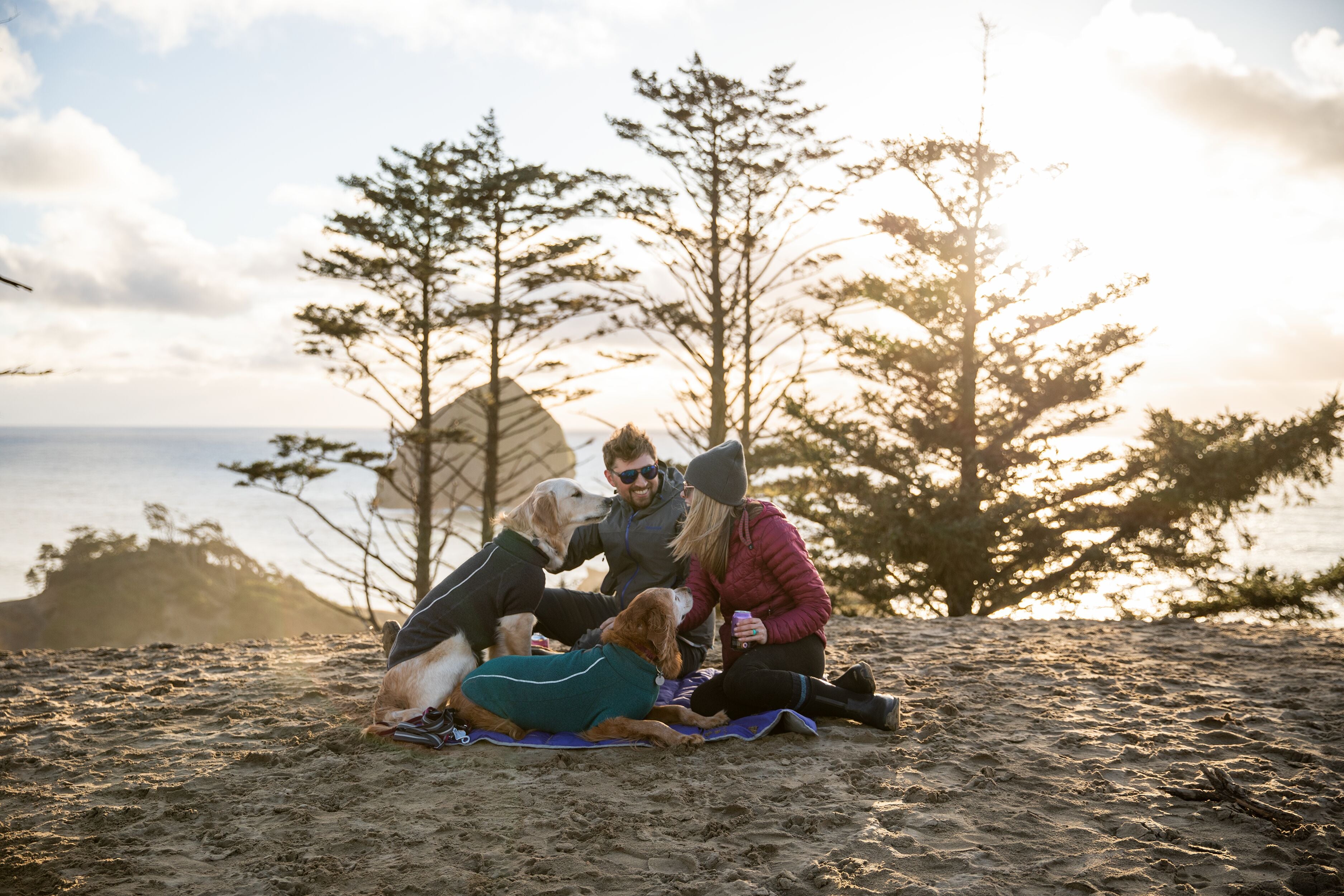A woman and man cuddle with their two golden retriever dogs at the Oregon Coast. 