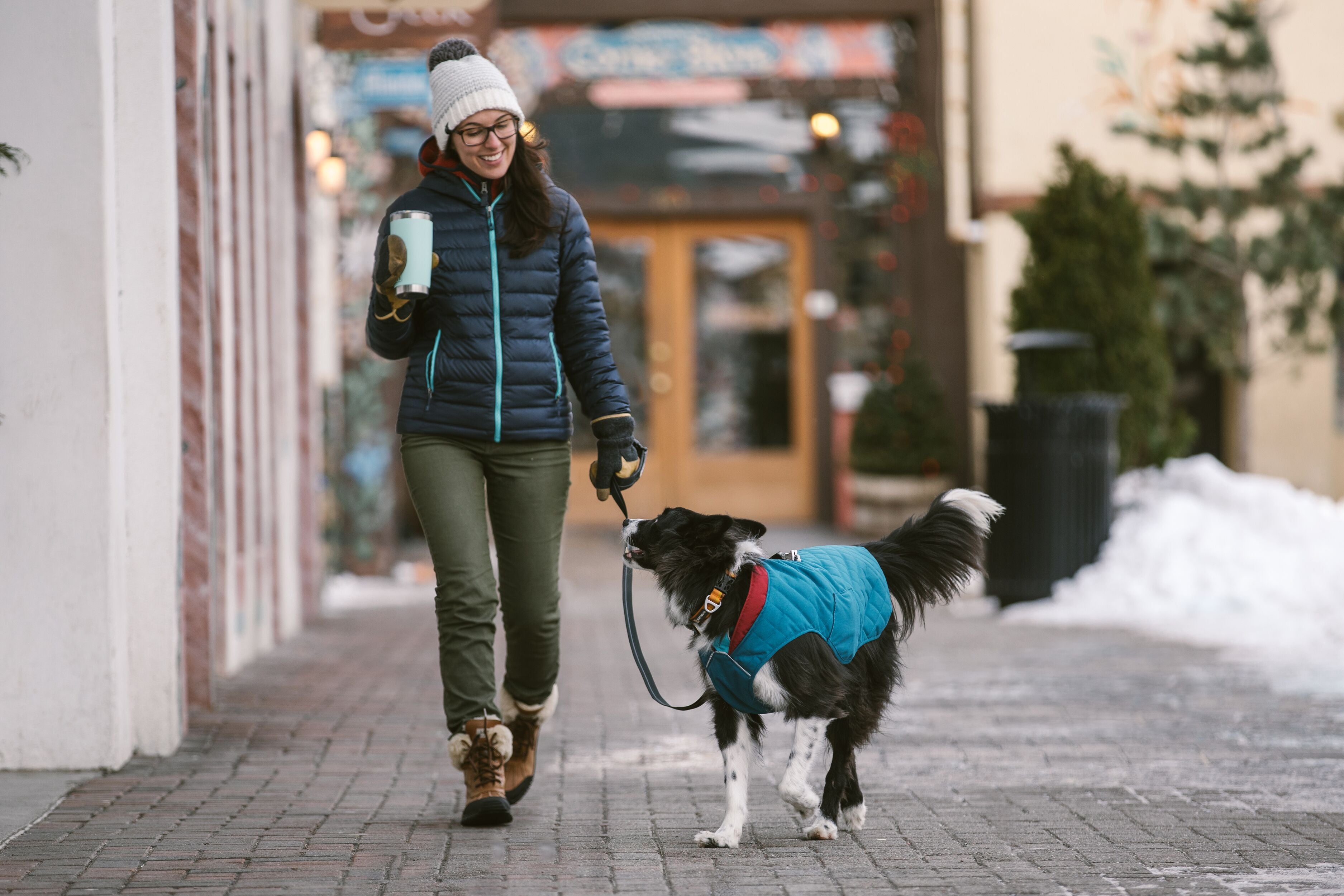 Dog in stumptown jacket walks with woman in city.
