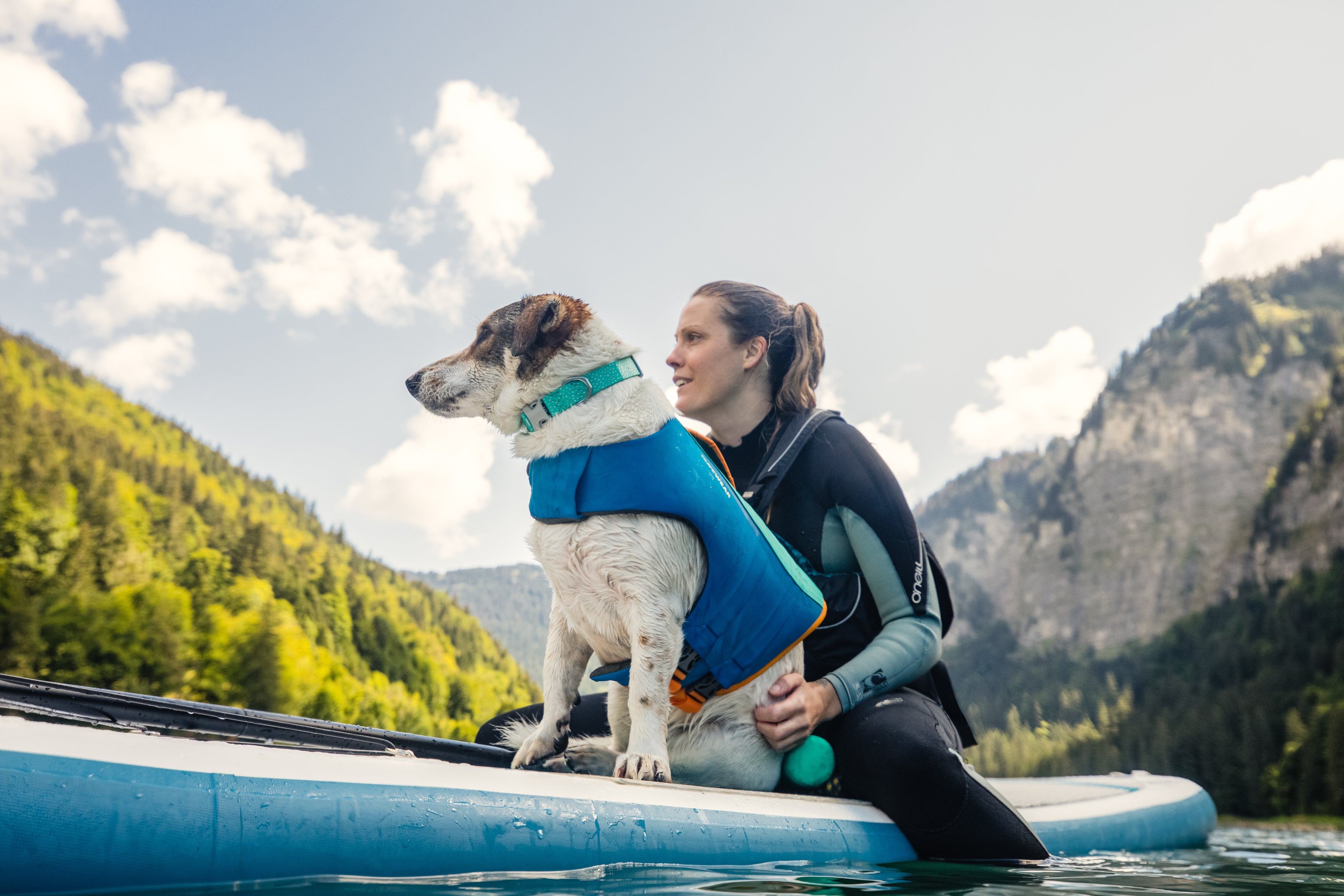 A woman sits with her dog on a paddle board in a lake. 