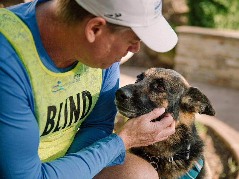 Man in yellow bib that reads blind with German shepherd guide dog sharing a moment.