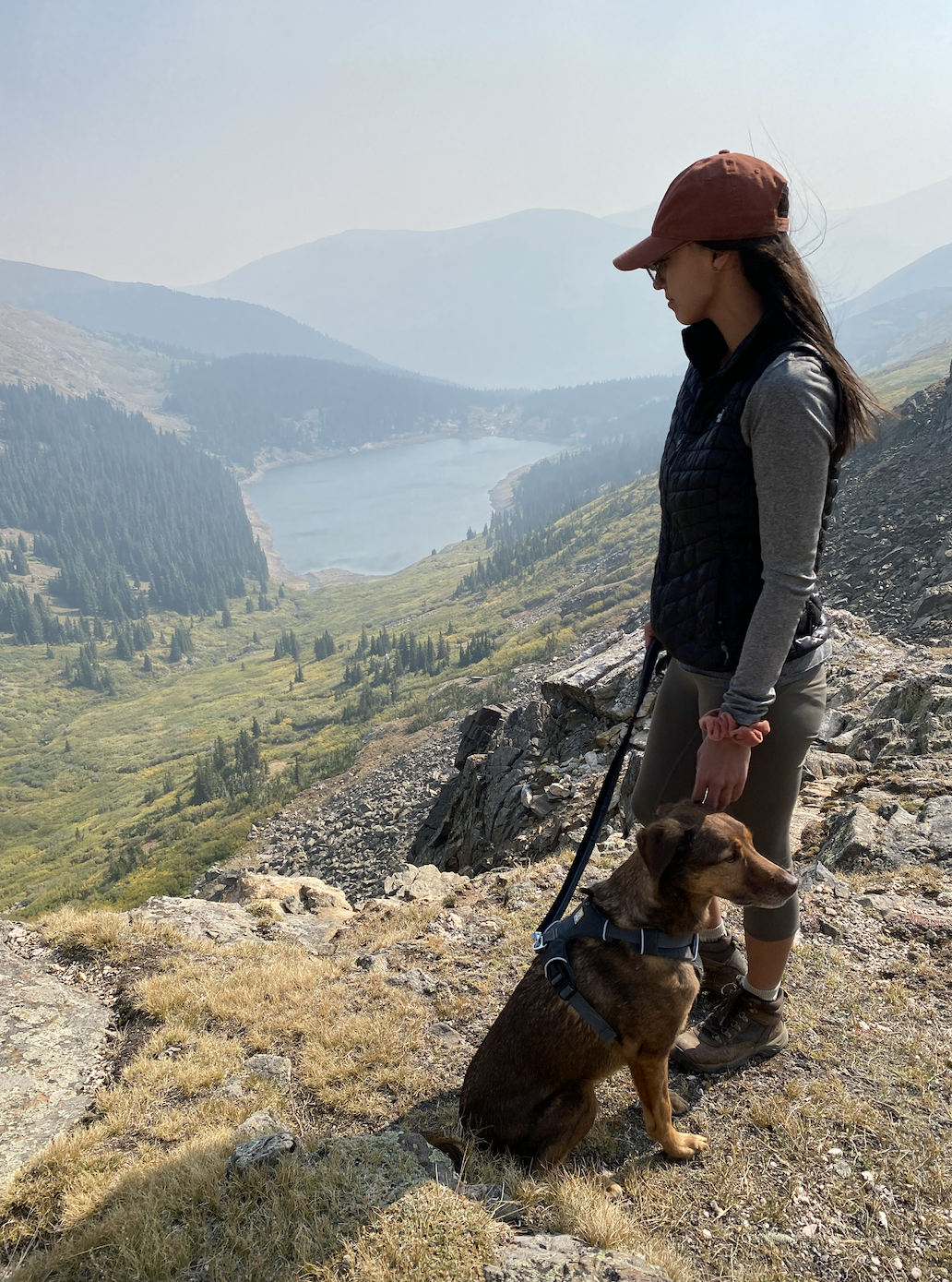 A woman stands with her dog on the top of a cliff while hiking. 
