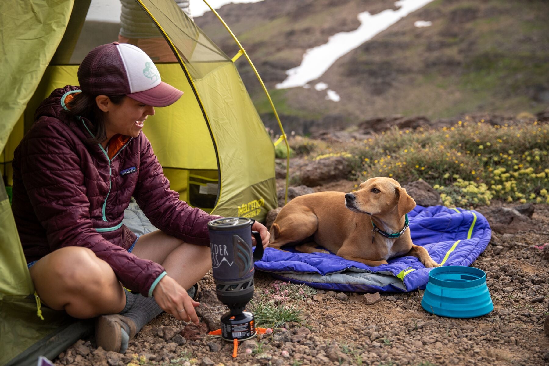 Human sitting in edge of tent cooking on her camp stove while dog sits next to her in Highlands dog sleeping bag.