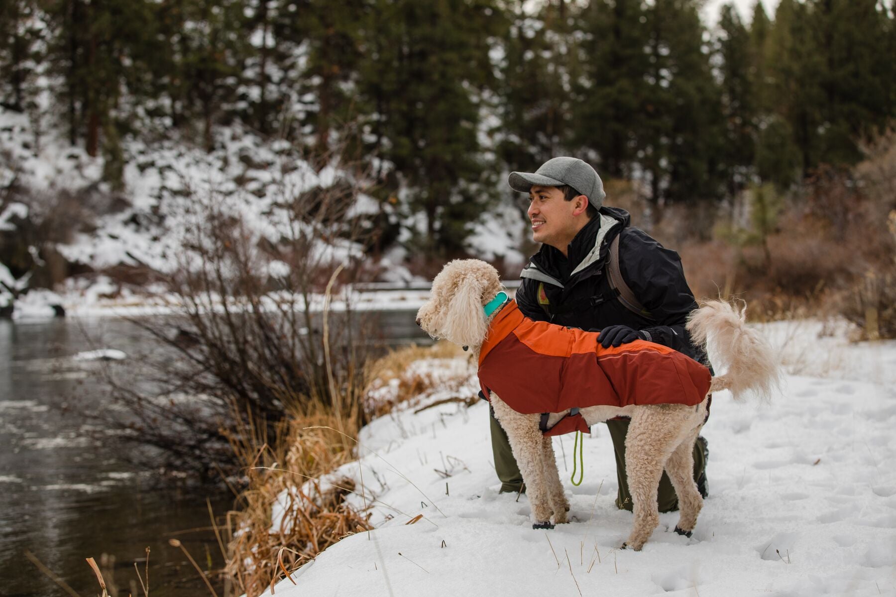 Man sits besides dog in Vert jacket on snowy trail.