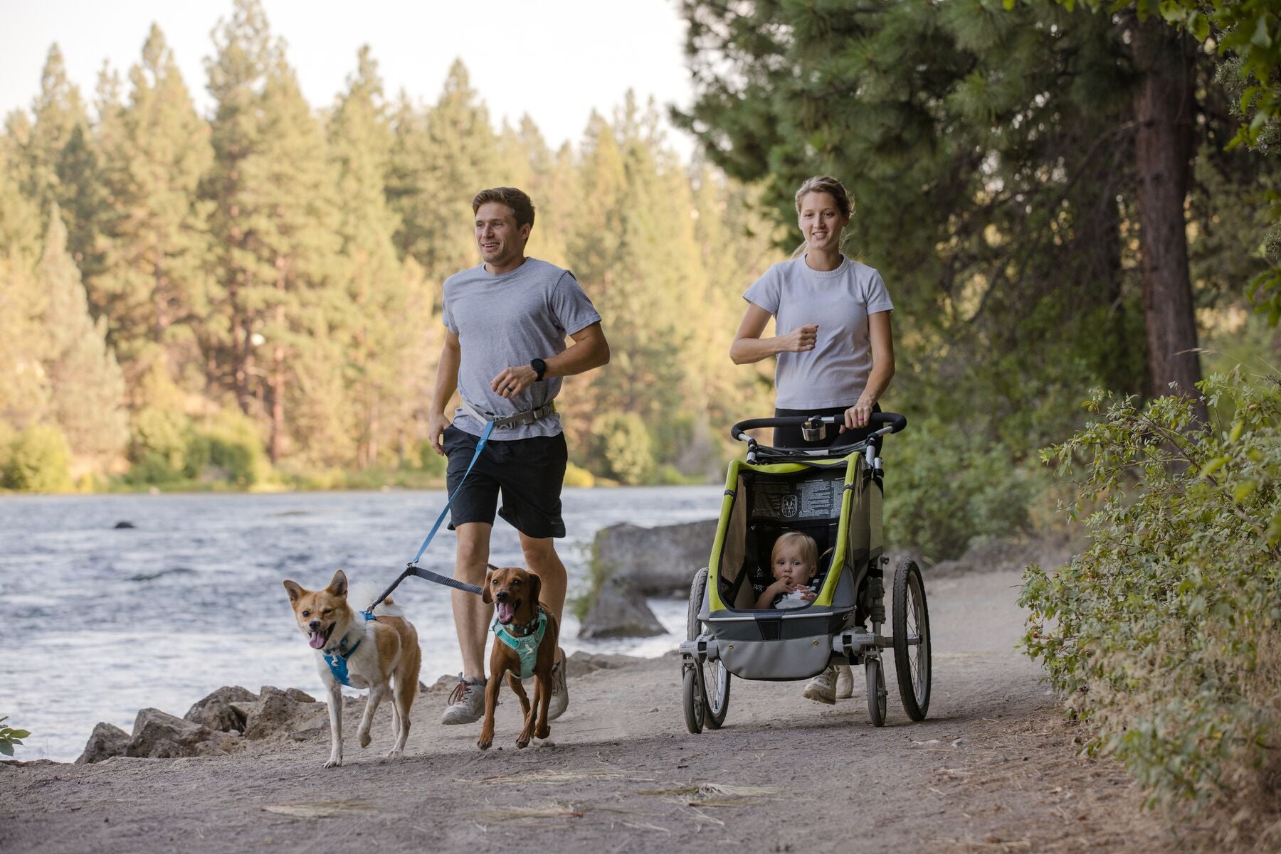 A man runs with two dogs on leash next to a woman running with a baby in a stroller