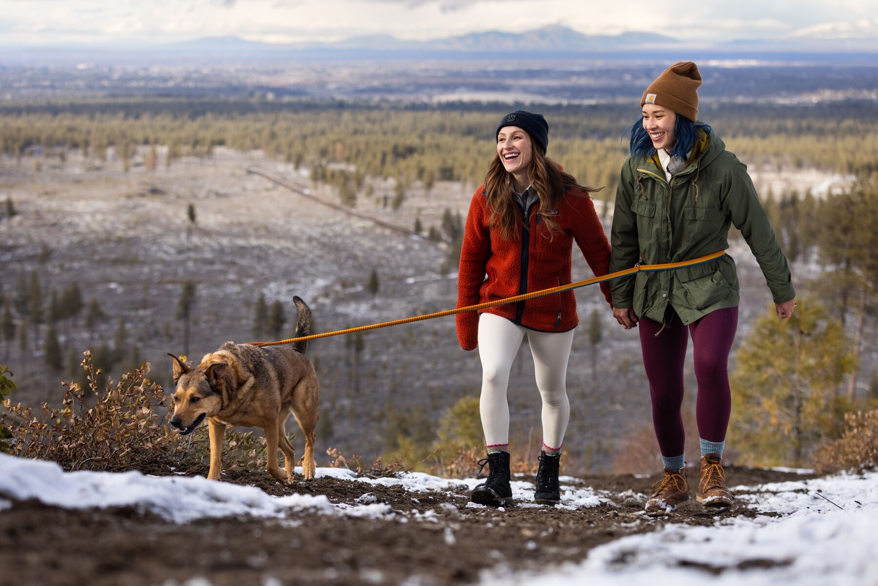 Two women hiking with their dog while their dog sniffs the trail