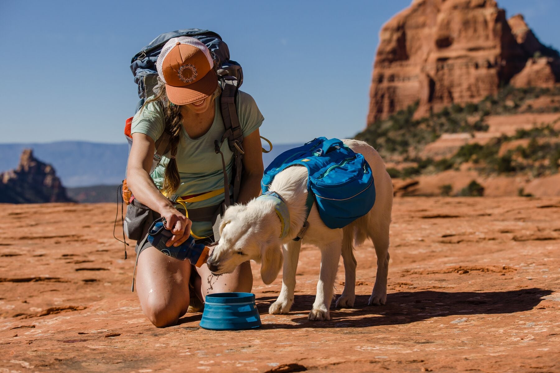 Woman with backpack kneeling down next to dog drinking water out of a bowl. 