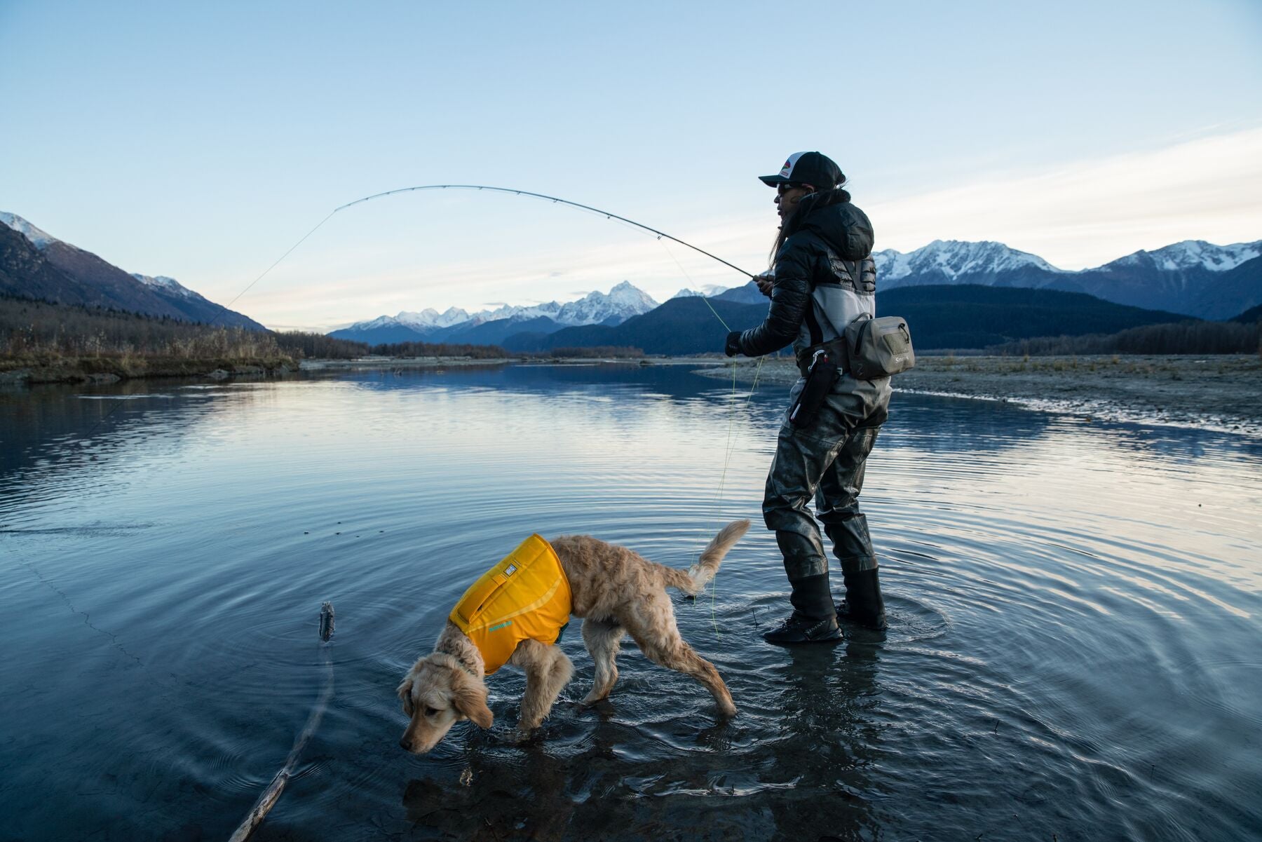 Woman fly fishing in river at base of Alaska mountains with puppy in float coat dog life jacket walking in water alongside her.