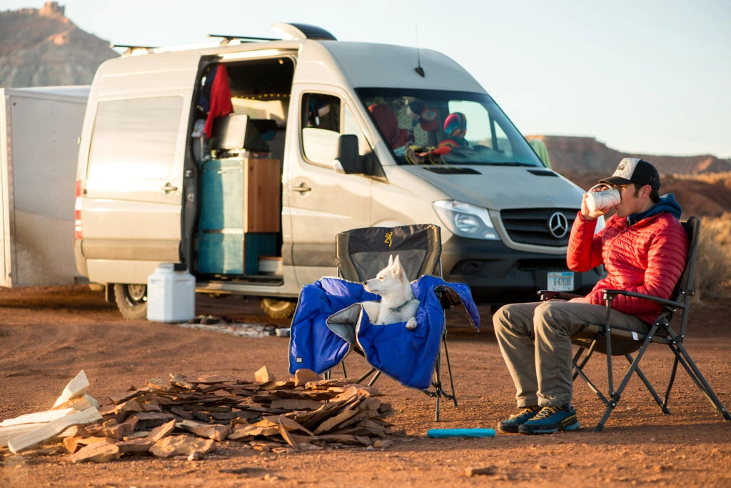 Dog wrapped in clear lake blanket sits in one camp chair while human sits in the other next to their van.