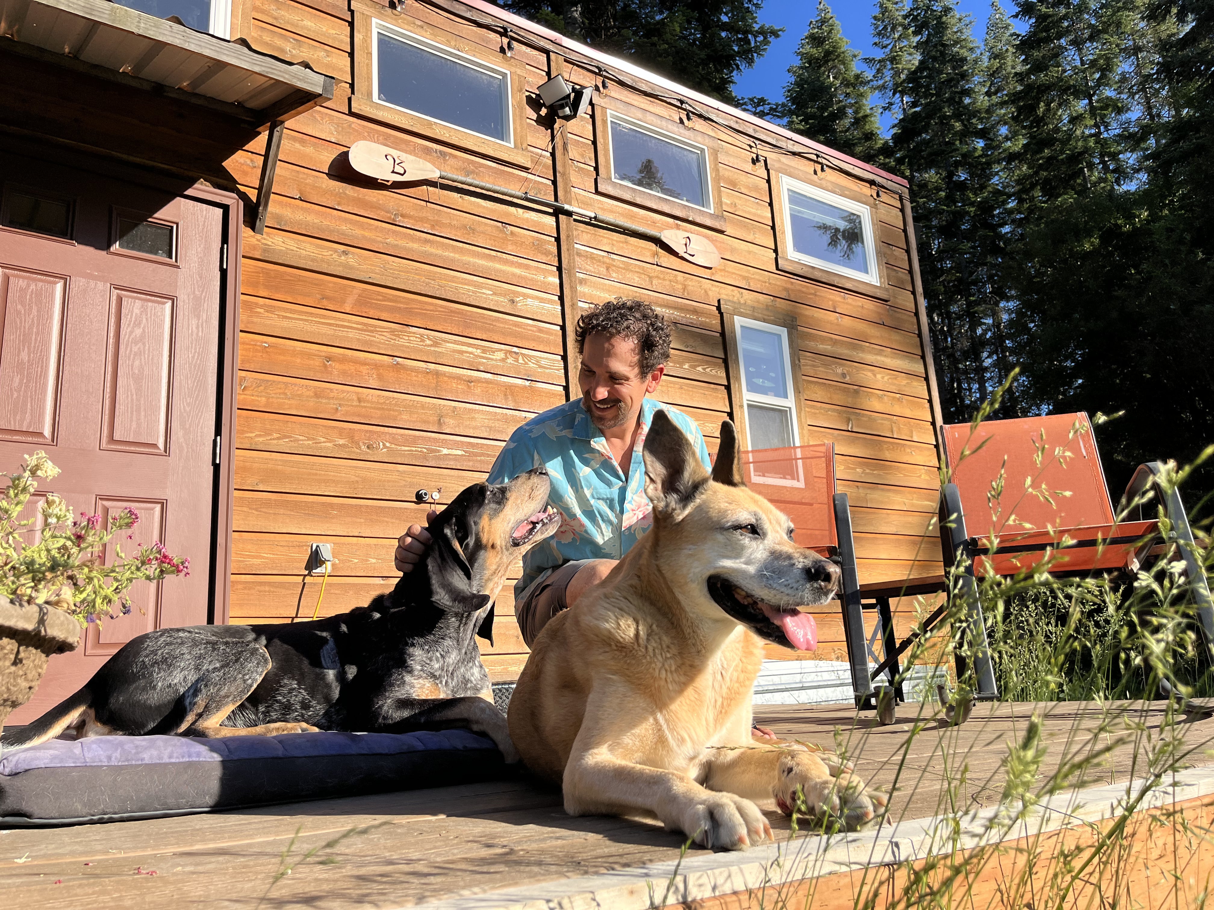 A man pets his two dogs outside their tiny home. 