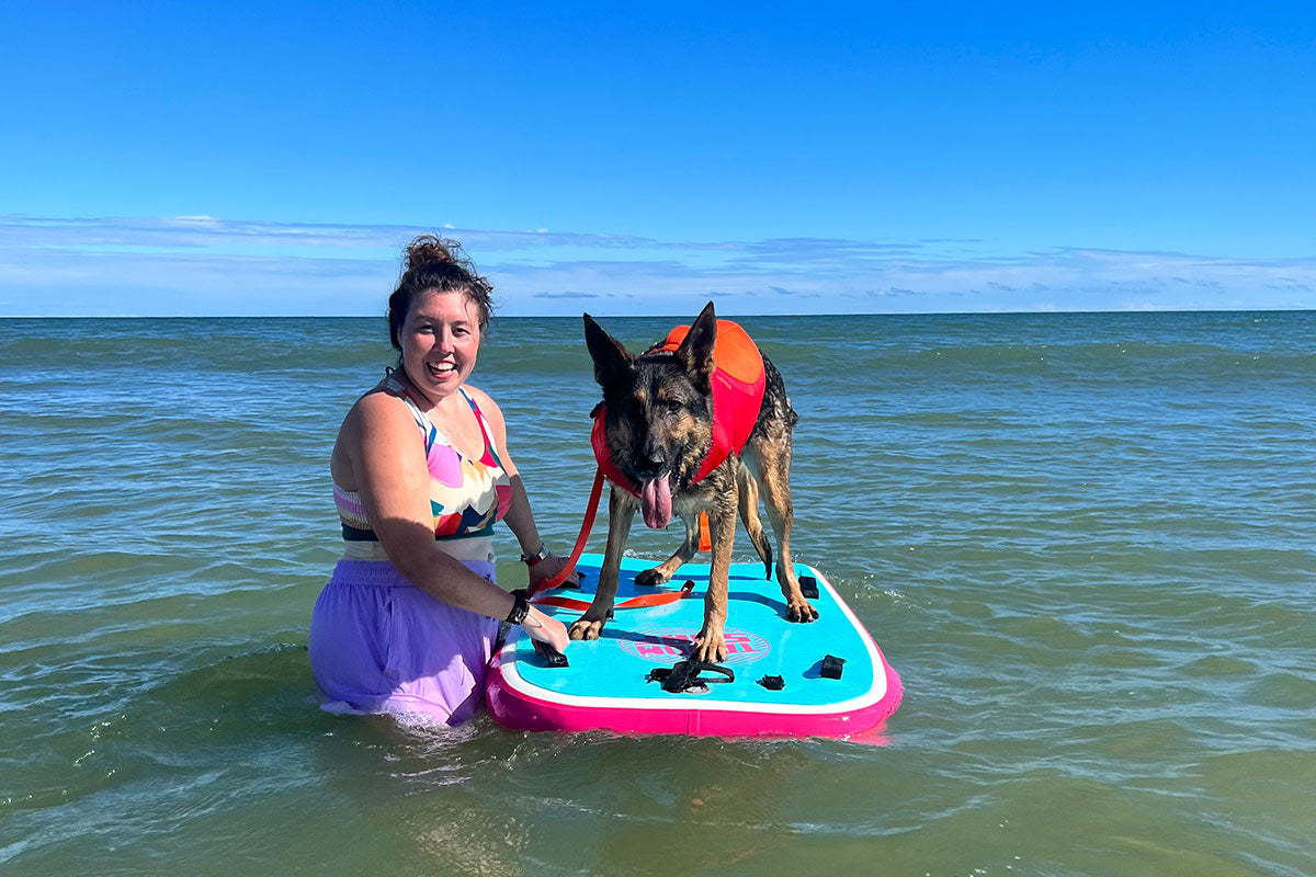 Zander wears a Float Coat atop a paddle board. Emma stands by his side in the water.