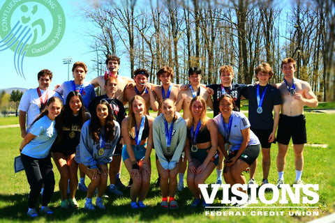 A group of cheerful students from Binghamton University's Health & Wellness Studies program pose outdoors after completing a triathlon. They are wearing medals around their necks, and there are clear blue skies and trees in the background. The students, a mix of genders and races, are smiling broadly, dressed in sportswear and shorts. Some are kneeling, while others stand behind them, all displaying a sense of accomplishment and camaraderie. Logos of "Confluence Running" and "Visions Federal Credit Union" are visible in the corners of the photo.