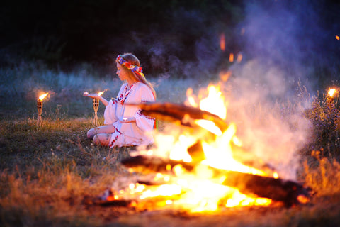 A bonfire burns in the foreground. In the background, a girl in Nordic dress and flower crown holds a lit candle.