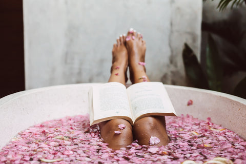 An open book lays on a pair of legs that rest on the ledge of a bath tub filled with pink flowers. 