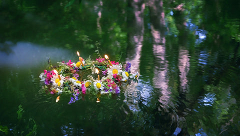 A flower crown with lit candles floats atop placid water, reflecting summer greenery.