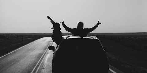 Silhouetted figures lean out from the sun roof and window of a car traveling down a long road