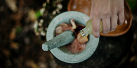 Rose petals in a mortar with pestle, and a hand adding something from a small glass bottle.