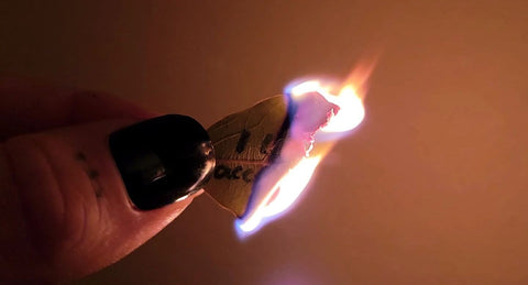 A hand with black nails holds a burning bay leaf with some partially obscured words in black ink still visible.