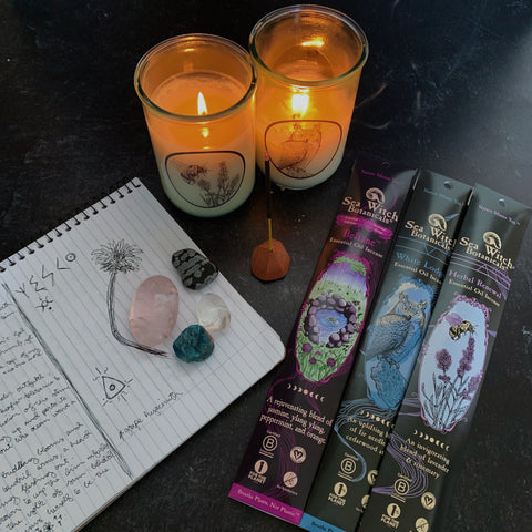 Two candles burning on a dark background with three incense packets, a stick of incense burning, a journal, and crystals