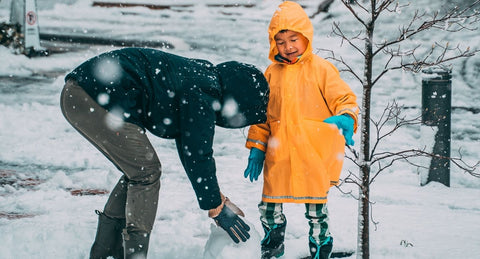 In a snowy scene, a parent leans down to form a snowman with their young child in bright yellow coat.