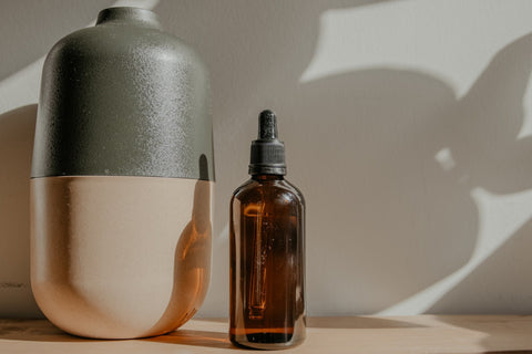 amber glass bottle topped with a dropper full of clear liquid next to a large essential oil diffuser, wood surface, plain background with shadows