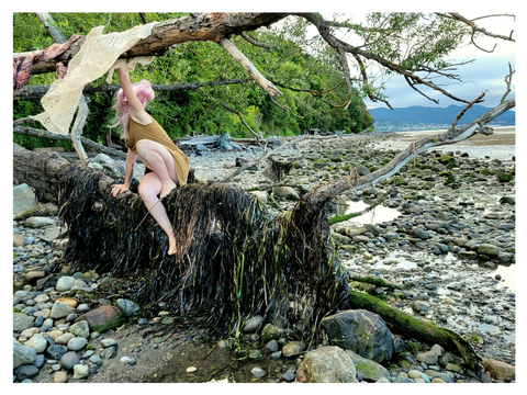 A woman with pink hair climbs along a horizontal tree over a rocky beach. photo by Madeleine (Peach) Ingridsdotter