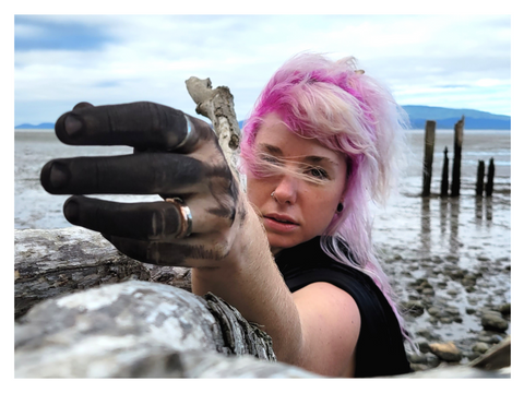 A wind-blown, pink-haired woman on a beach rests her arm on a tree branch, reaching toward the camera. Her fingers are blackened with charcoal. photo by Madeleine (Peach) Ingridsdotter