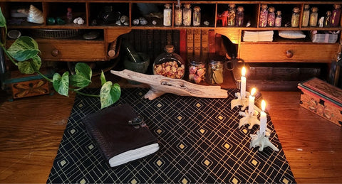 Upon an old desk with small shelves lined with jars of different things, a black and gold cloth is set with a journal, incense, and lit candles.