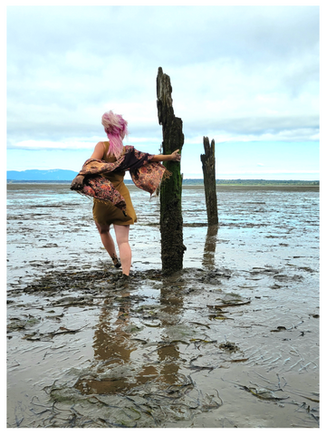 Sea Witch Botanicals founder Alesia Hall dances barefoot in the muddy low tide and high wind on the Pacific coast. She has pink hair and a mustard yellow dress. 