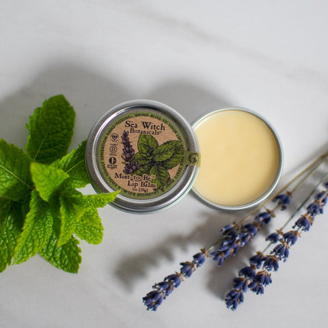 Opened tin of Mint-to-Be Lip Balm with fresh mint and lavender on white background.