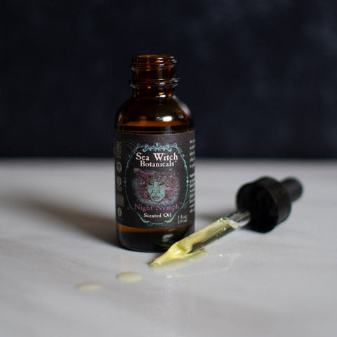 Opened bottle of Night Nymph Scented Oil with dropper on marble countertop, dark background.