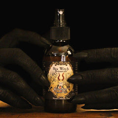 Soot-black hands with claw-like nails around a bottle of limited edition Krampus essential oil scented veil and room spray.