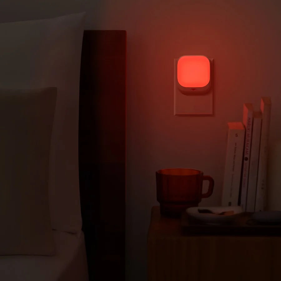 A red night light plugged into a wall near a bed and a bedside table with a mug and books.