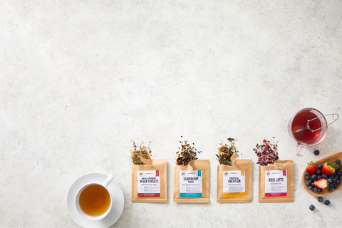 tea sampler box with 4 sample packets open with loose leaf tea pouring out; freshly brewed tea and fresh berries on side; white marble background