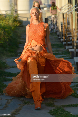 Model walking the runway with a blonde buzz cut wearing an orange jumpsuit with lots of ruffle detailing and a long train that flows behind them over their pants 