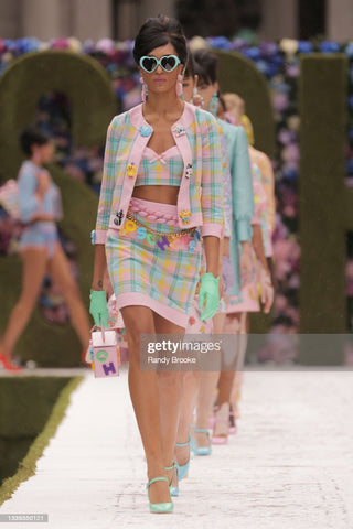 Runway model in a two piece set that is colorful pastel plaid pattern with a skirt and a blazer that match. She is wearing sunglasses and a belt made of colorful block letters that spell "Moschino"