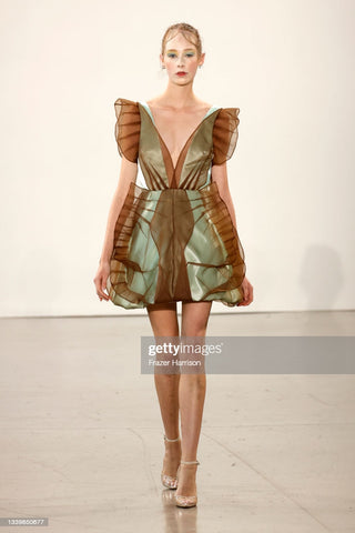 Runway look, model wearing a green shiny short aline dress with sheer brown detailing that extends past the shoulders and stretches. It looks like it is inspired by a butterfly.