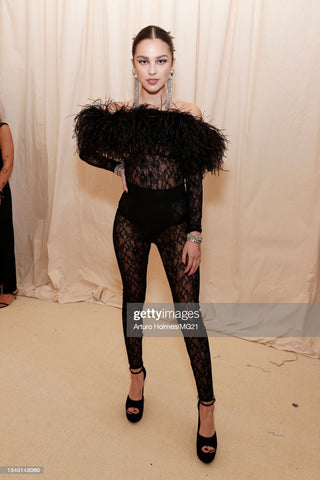 Singer and pop icon olivia rodrogo in a black all-lace jupsuit that has black feather off the shoulder details covering the breast area. She is wearing long diamond earrings and extremely tall black platform heels 