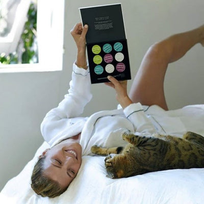 a woman lying down on a bed next to her cat, trying to decide which of the best clay masks she wants to use