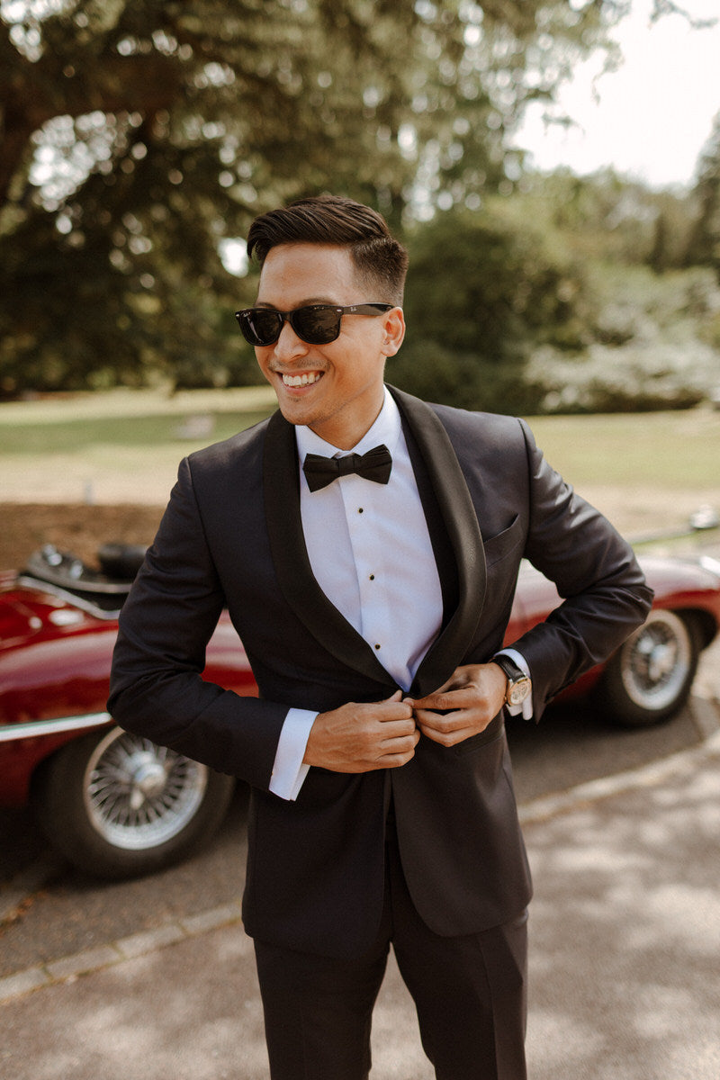 Groom patrick wears a bespoke three piece suit and sunglasses, he stands in front of a vintage car.