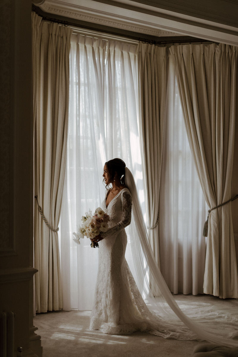 Bride Charlotte stands at a window in her Chantilly lace wedding dress and veil, holding a bouquet.