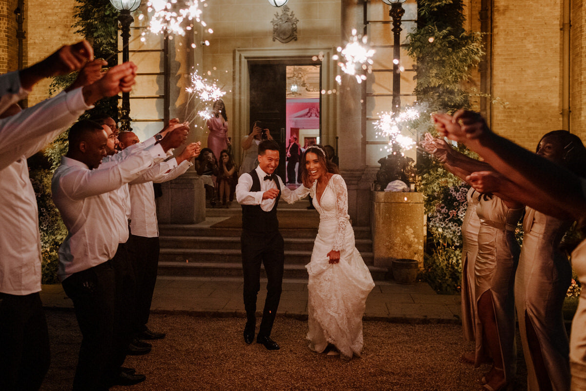 Bride and groom exit their wedding reception venue, flanked by sparklers held by their guests.