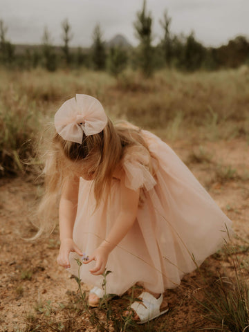 A young girl picks a flower, wearing our champagne Isla flower girl dress and champagne tulle bow hair clip.