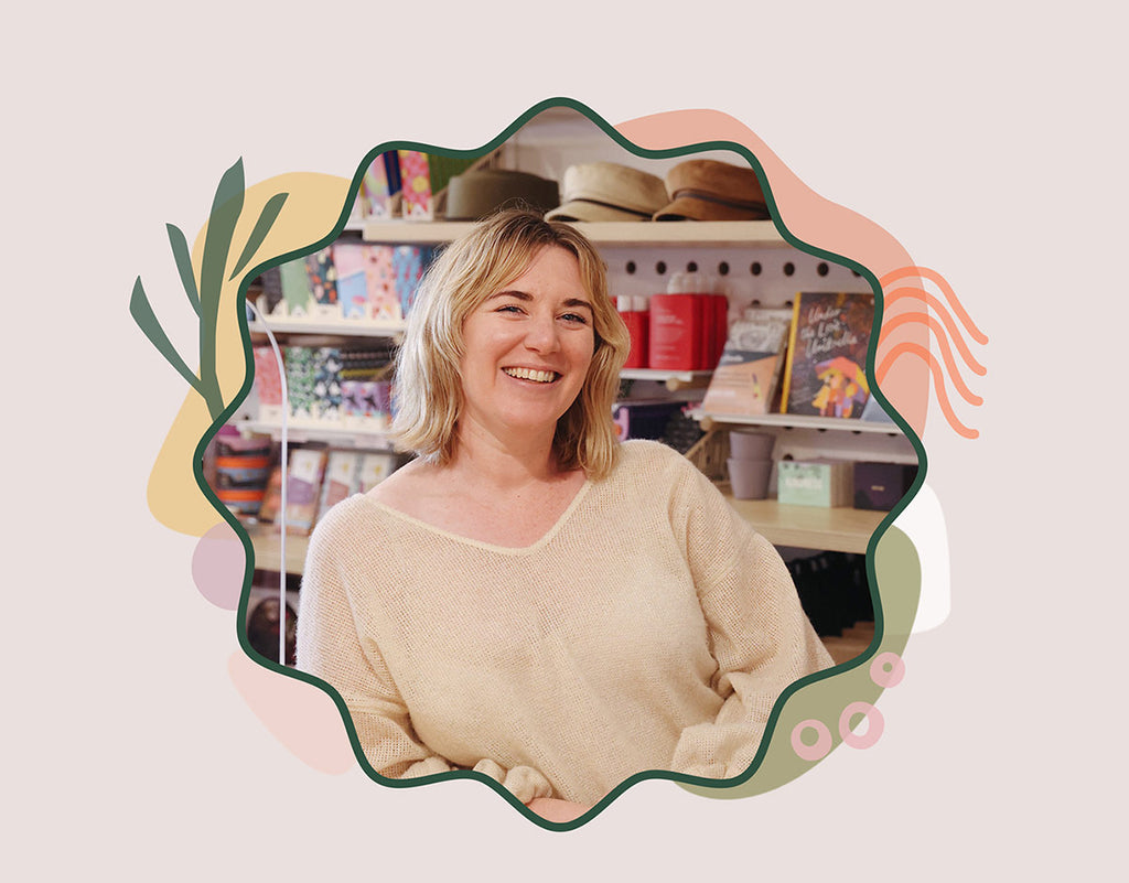 Portrait of Kylie, owner of A Quirk of Fate in the shop, smiling to the camera. Kylie wears a cream wool top, she has blonde hair and fair skin; and is surrounded by colourful shelves of product in-store.